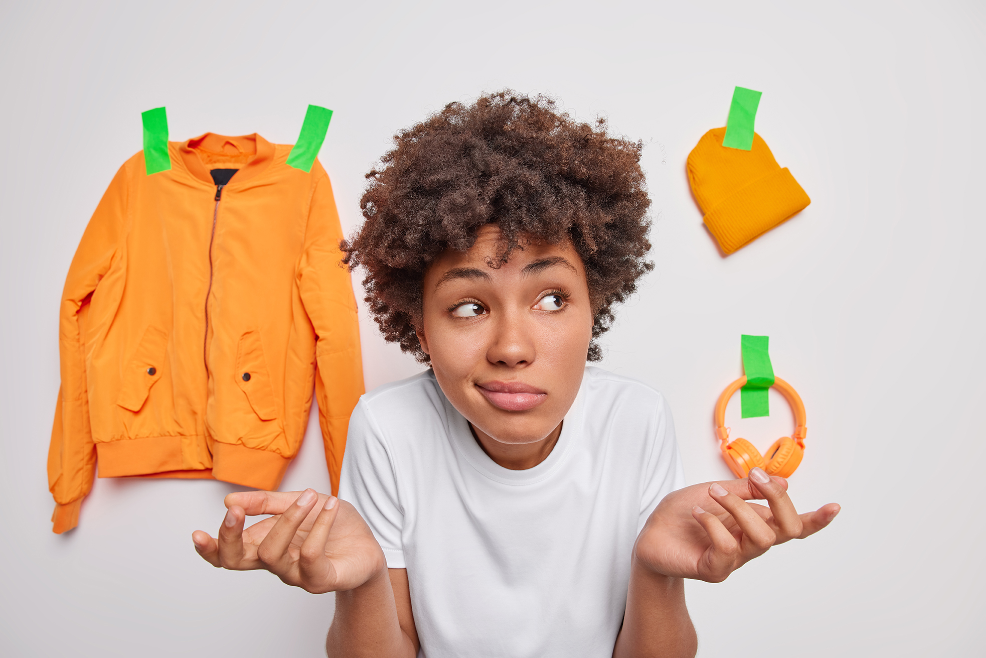 Clueless hesitant curly haired woman shrugs shoulders has questioned expression focused away poses against white background with plastered items doesnt know what to choose faces difficult choice